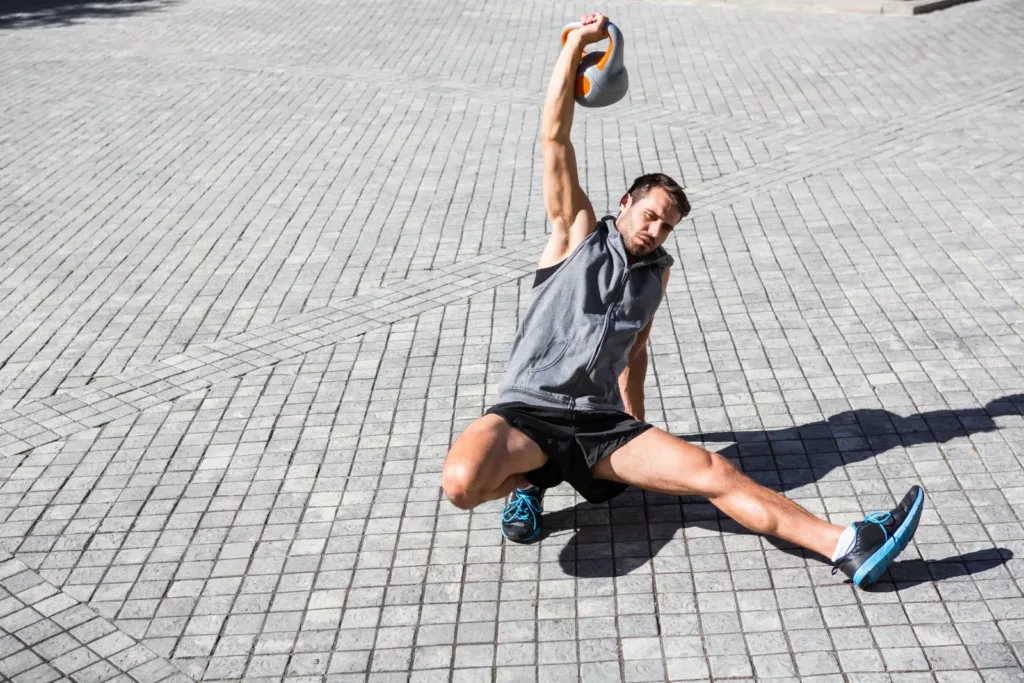 Man in a half-kneeling position on paving with a kettlebell locked out overhead during a Turkish get-up