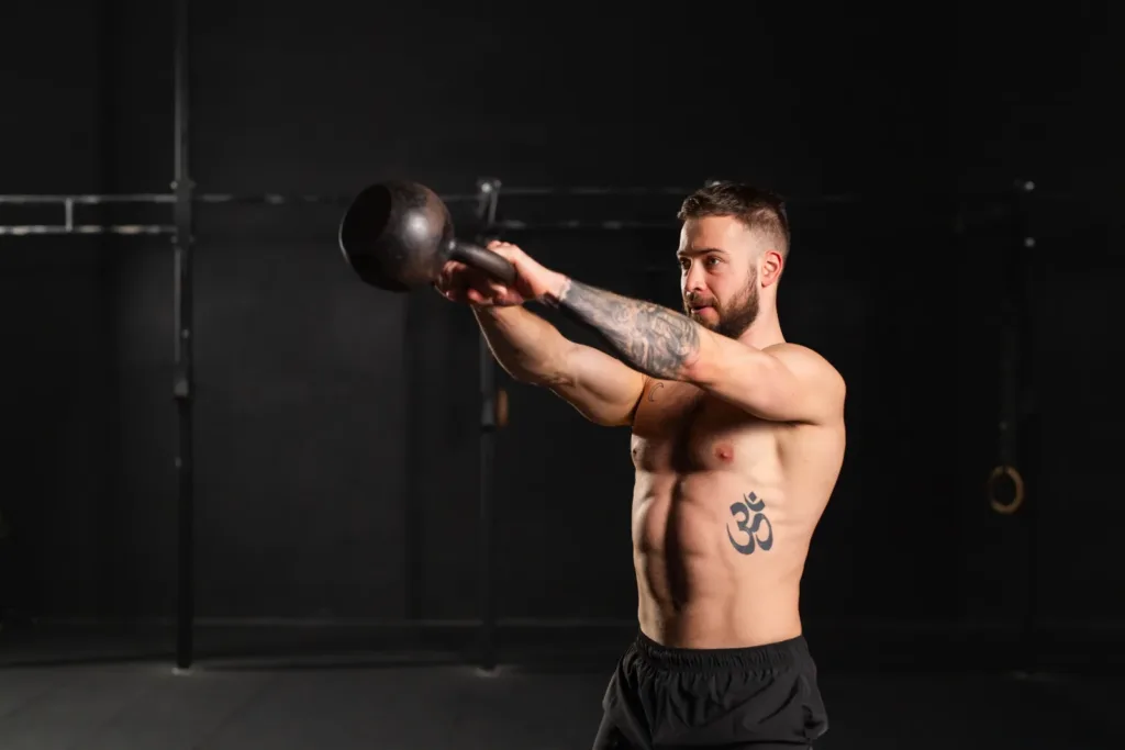 Shirtless man at the top of a kettlebell swing, arms straight at chest height