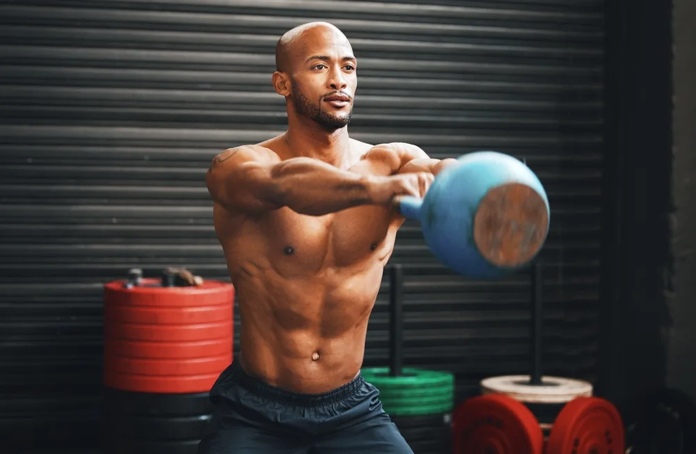 Shirtless man swinging a blue kettlebell to chest height in a gym