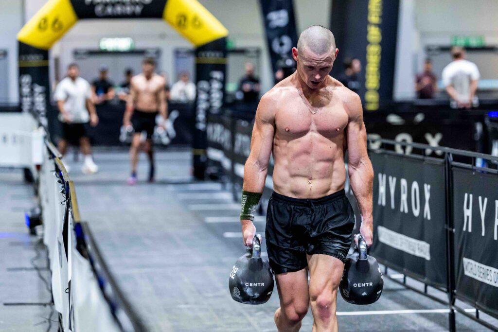 A bald, muscular HYROX competitor carrying two kettlebells during the farmers carry station