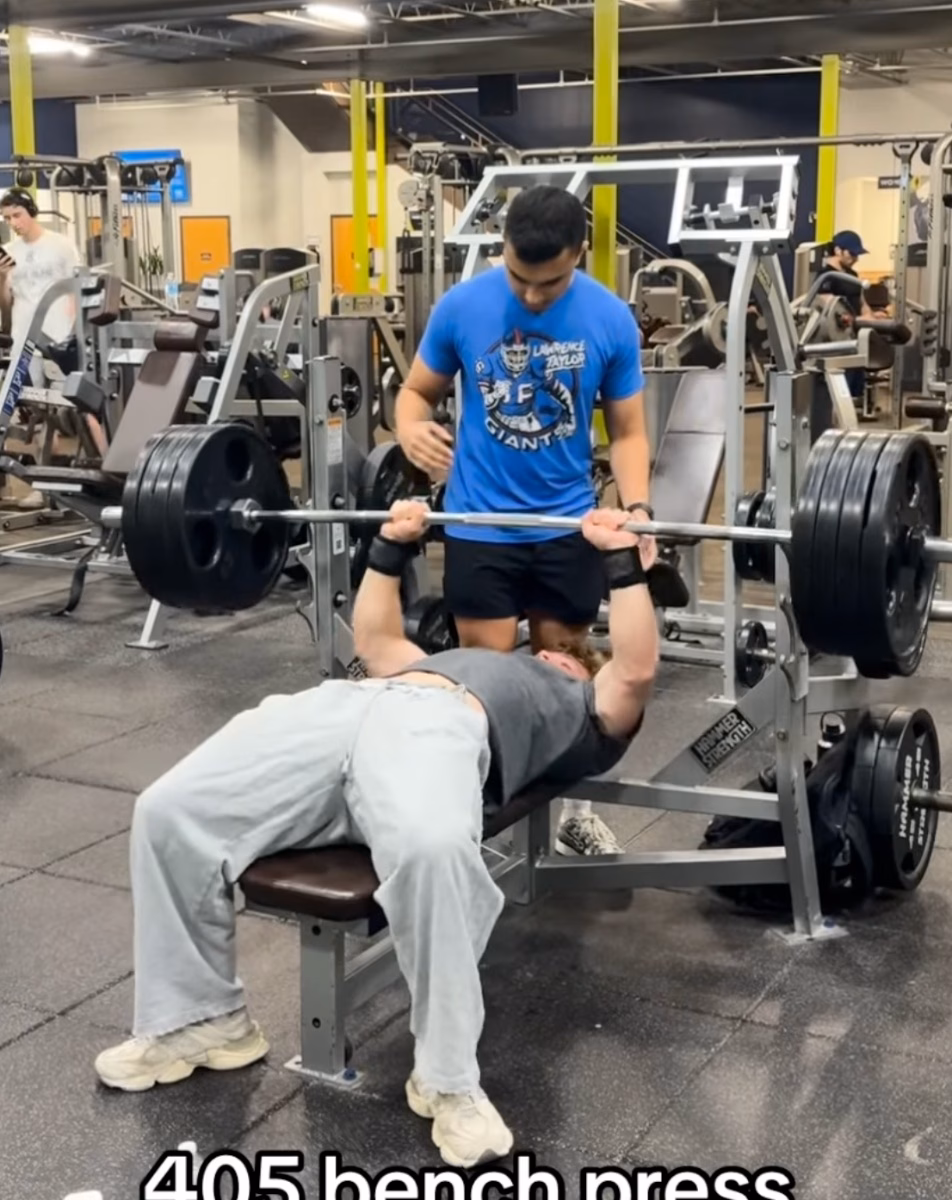 A lifter bench presses a heavily loaded barbell in a commercial gym as a spotter in a blue T-shirt stands behind him, with on-screen text reading 405 bench press