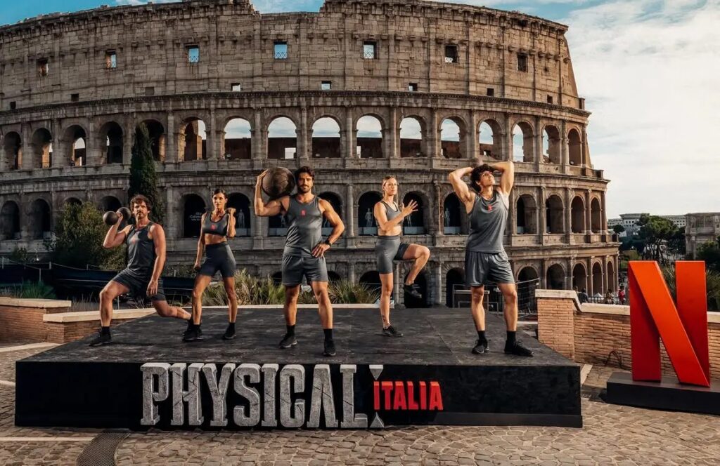 Five Physical 100: Italy contestants in grey team kit pose with a kettlebell and medicine ball on a Physical Italia stage in front of the Colosseum in Rome, beside a Netflix logo