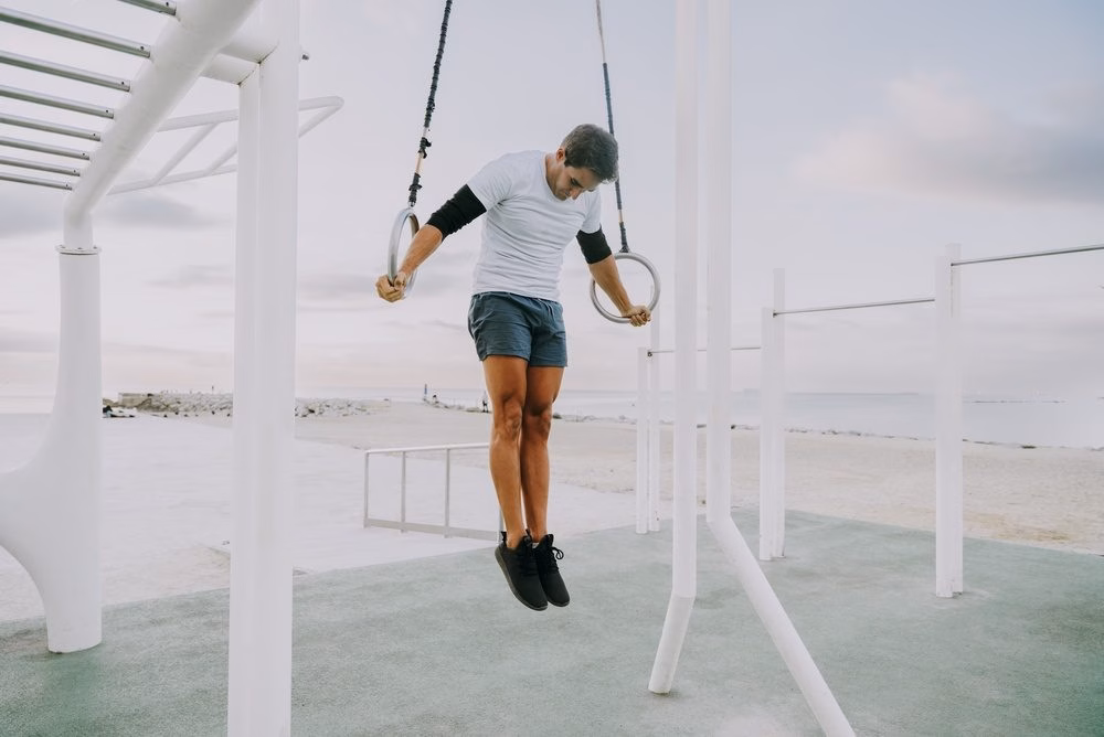 Man pulling through a ring muscle-up at an outdoor beachside calisthenics park.