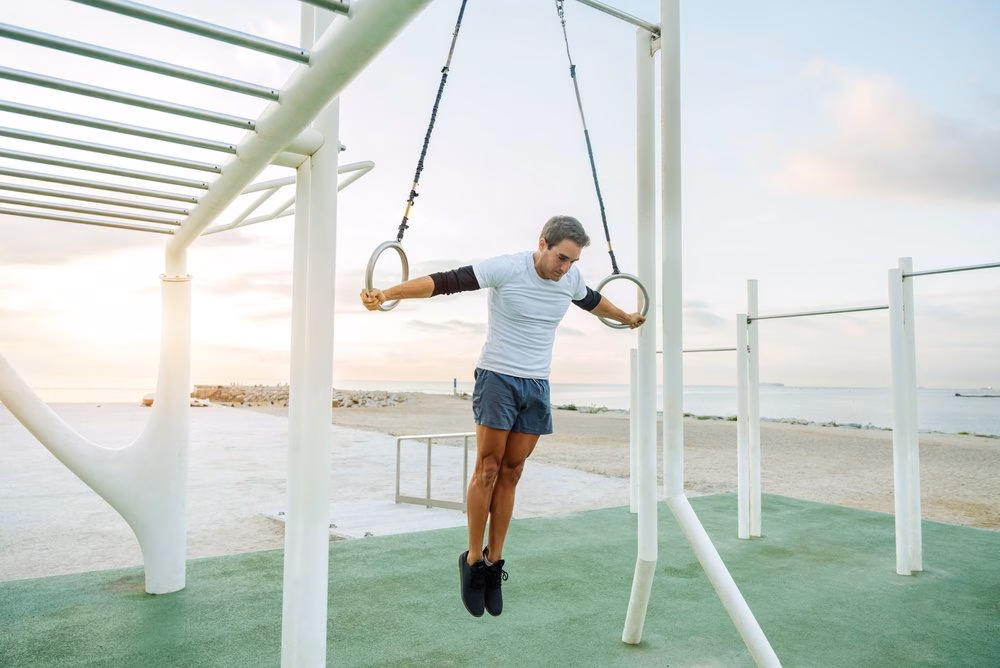 Man performing a support hold on gymnastics rings at an outdoor fitness park, arms extended to the sides.