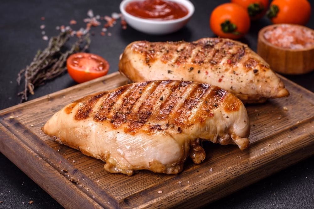 Two grilled chicken breasts on a wooden board with tomatoes and salt, one of the highest-protein foods per 100g