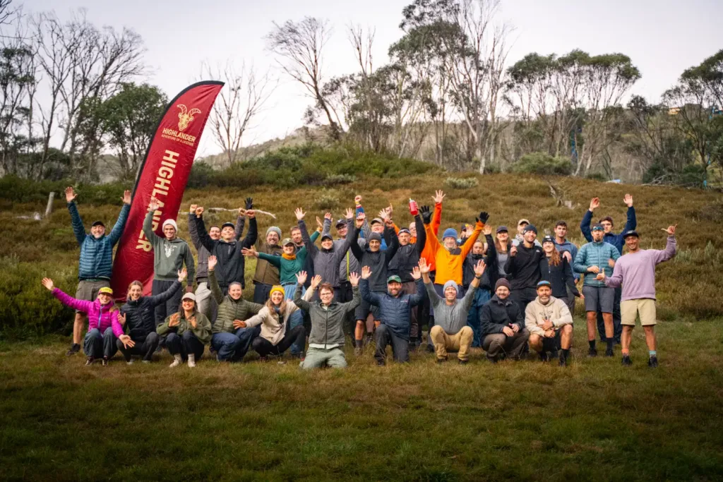 A large group of hikers with arms raised standing next to a red HIGHLANDER Adventure flag