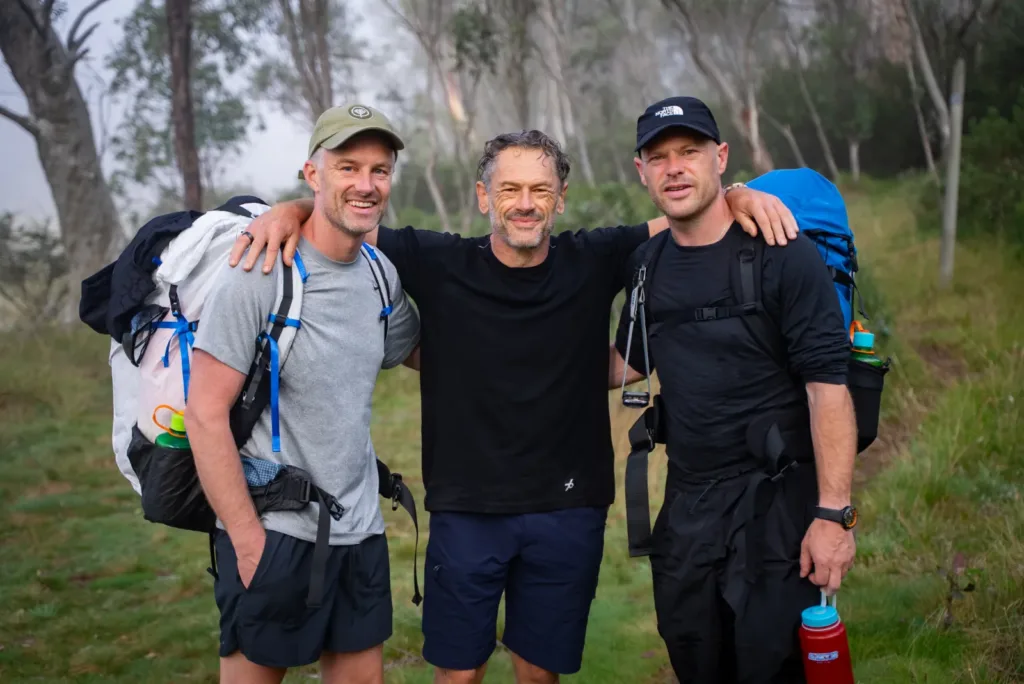 Three hikers with backpacks smiling and embracing after completing a HIGHLANDER hike