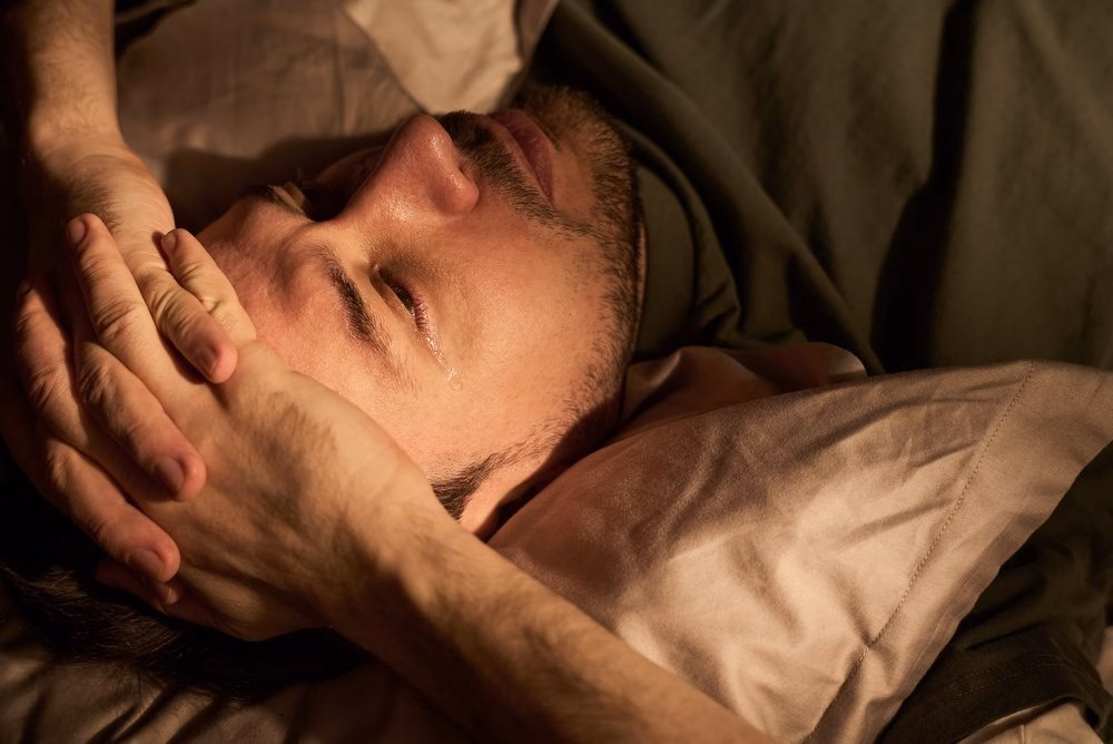 Man lying in bed at night with his eyes closed, looking distressed and unable to switch off