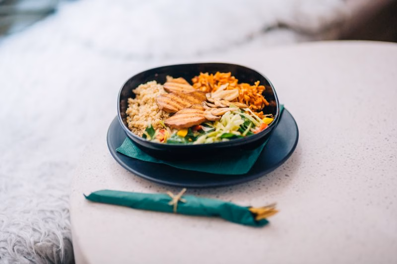 Black bowl of quinoa, grilled sliced protein, shredded carrot and mixed vegetables on a green napkin