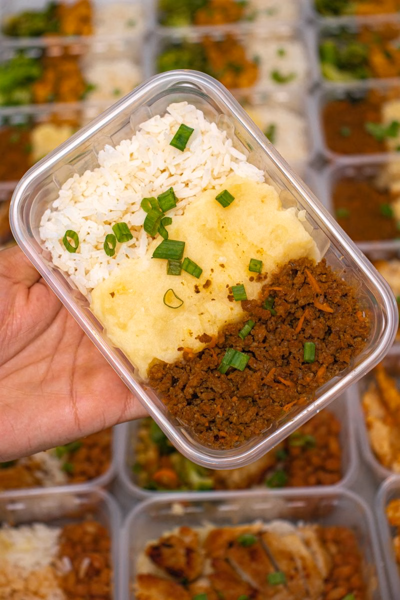 Hand holding a meal prep container of white rice, mashed potato and seasoned ground beef topped with spring onion