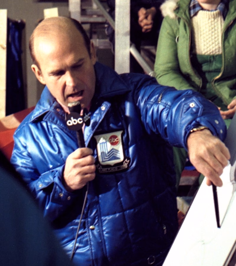 Dick Button in an ABC jacket commentating into an ABC microphone at an ice rink