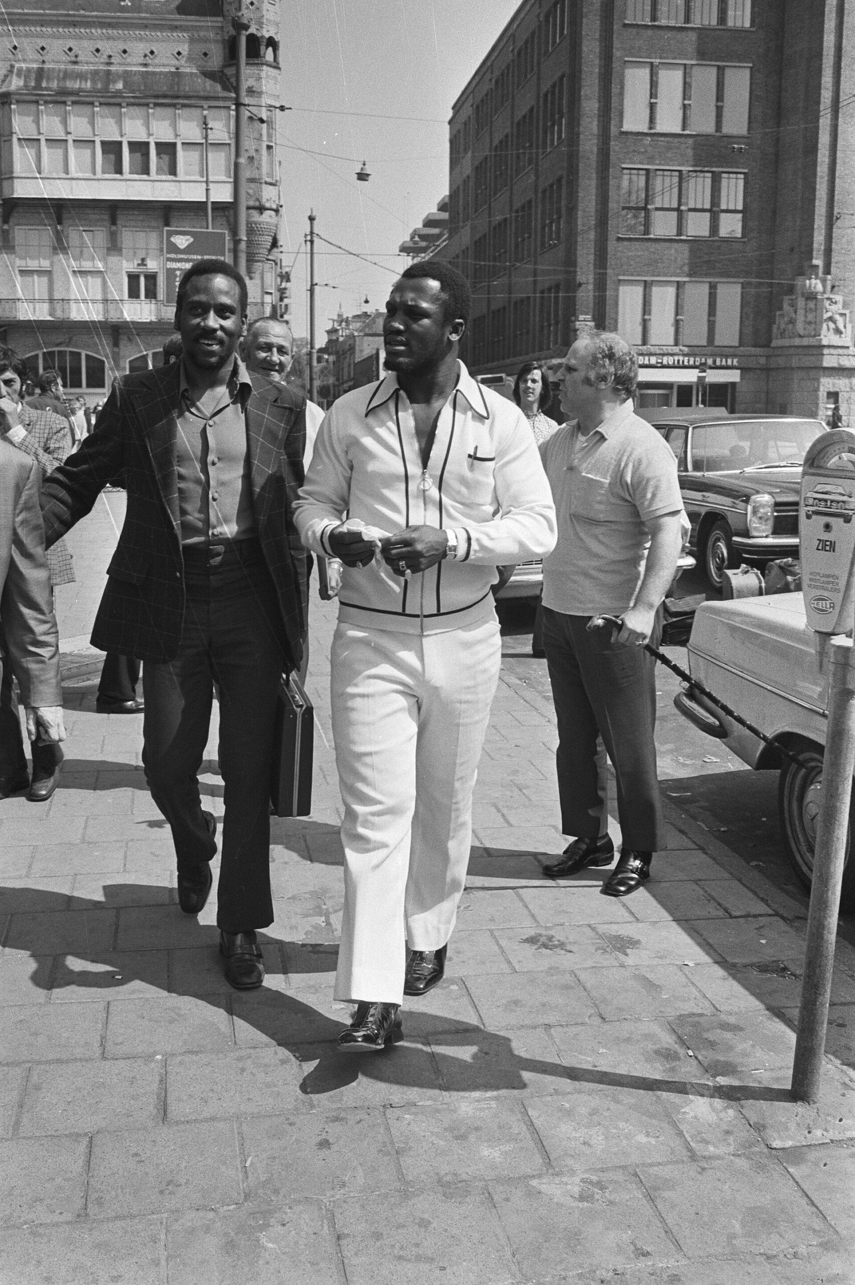 Black-and-white photo of boxer Joe Frazier in a white tracksuit walking down an Amsterdam street