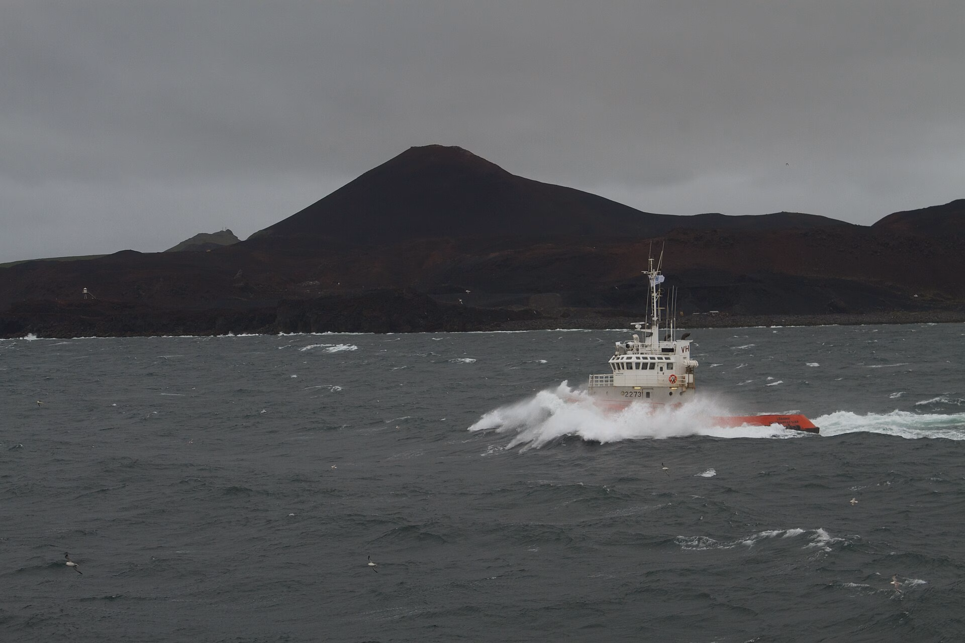 A small boat ploughing through grey, choppy sea off Heimaey, with dark volcanic hills behind under heavy cloud