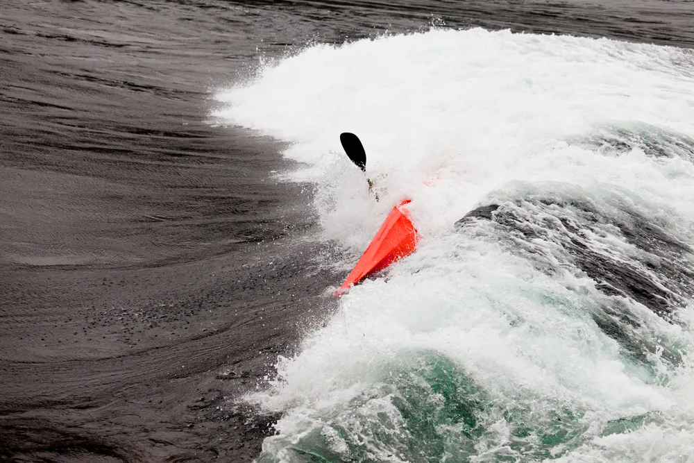 A kayaker in a red kayak is swallowed by a breaking wave in grey water