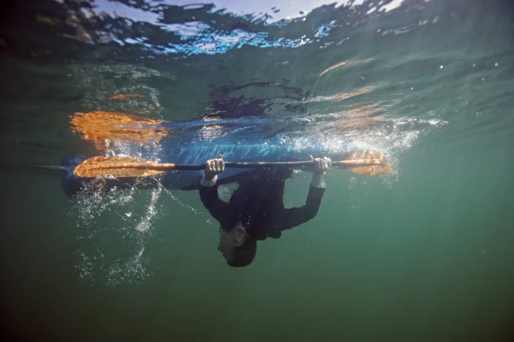 Underwater view of a capsized kayaker hanging upside down beneath a kayak, still gripping the paddle