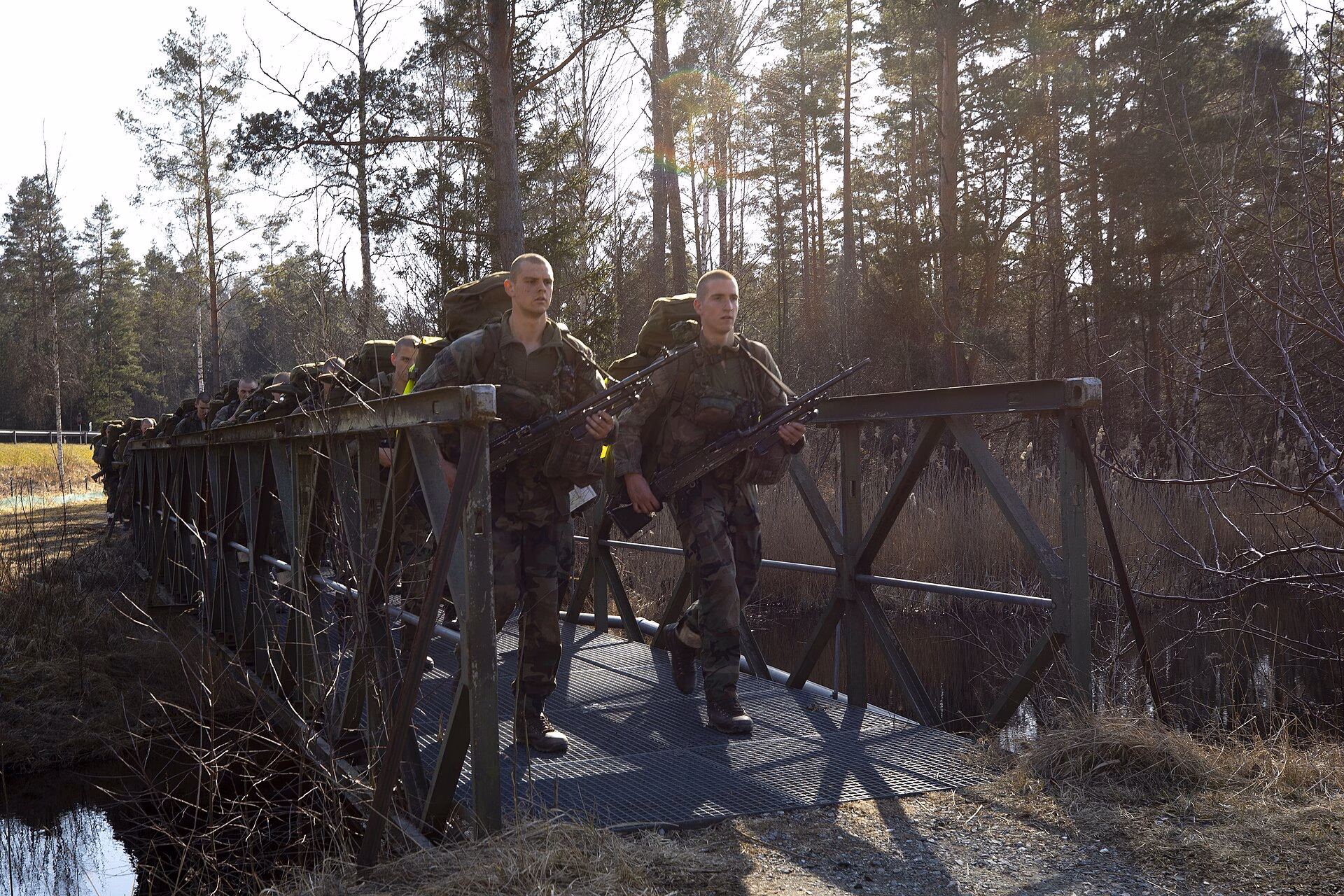 A line of Dutch Marine recruits with heavy packs and rifles crosses a metal footbridge in a pine forest