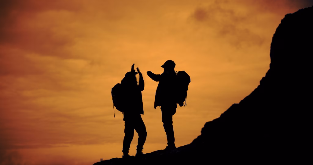 Two people wearing backpacks give each other a high five, silhouetted on a rocky ridge against an orange sunset sky